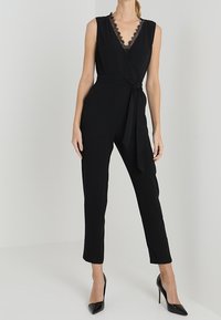 Tuta nera con scollatura a V, bordo in pizzo e vita con cintura. Vestibilità sartoriale con gambe affusolate, abbinata a tacchi alti neri.