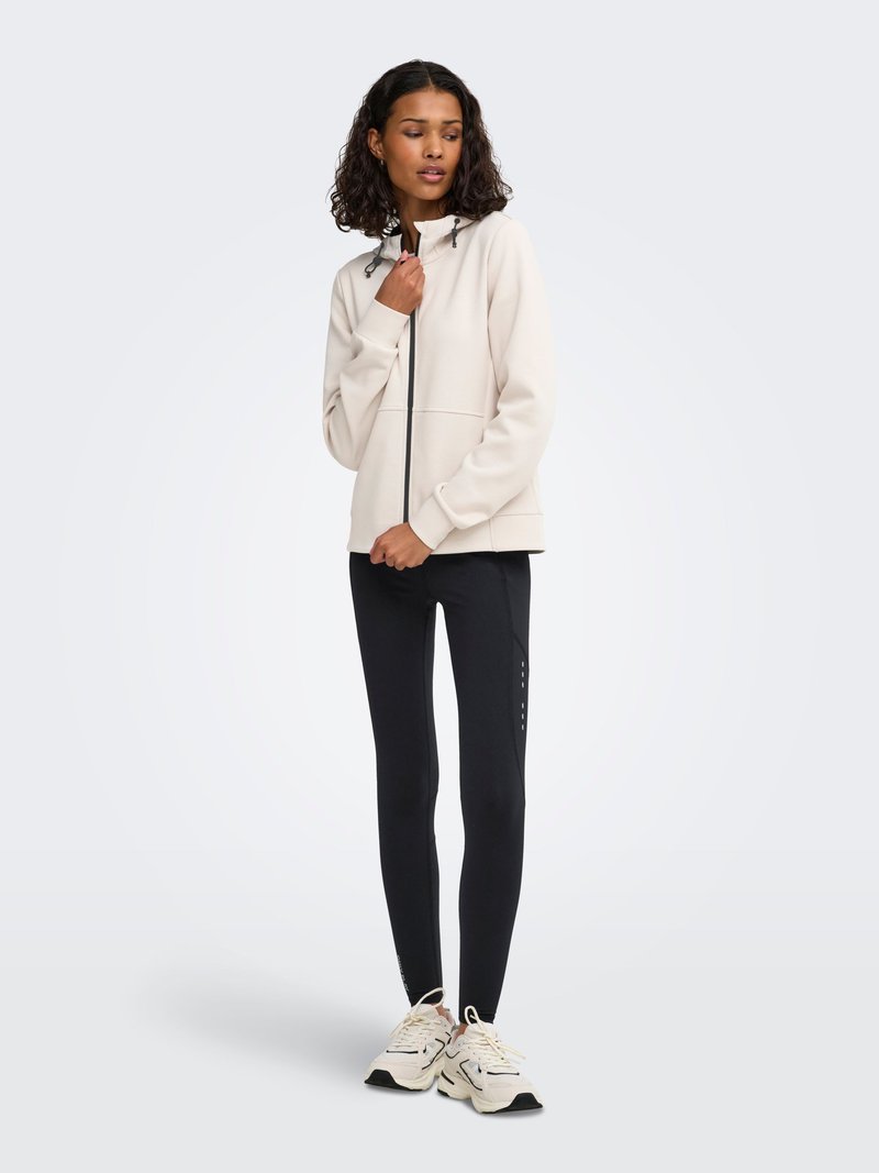 Femme debout portant une veste zippée beige clair, des leggings noirs avec un détail latéral et des chaussures de sport de couleur claire sur un fond uni.