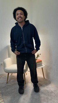 Νέος άνδρας με σγουρά μαλλιά, φορέας μπλε σκούρας ζακέτας με φερμουάρ και μαύρο παντελόνι, στέκεται σε ένα δωμάτιο με μοκέτα, όπου βρίσκεται μια άσπρη πολυθρόνα και ένα μικρό τραπέζι.