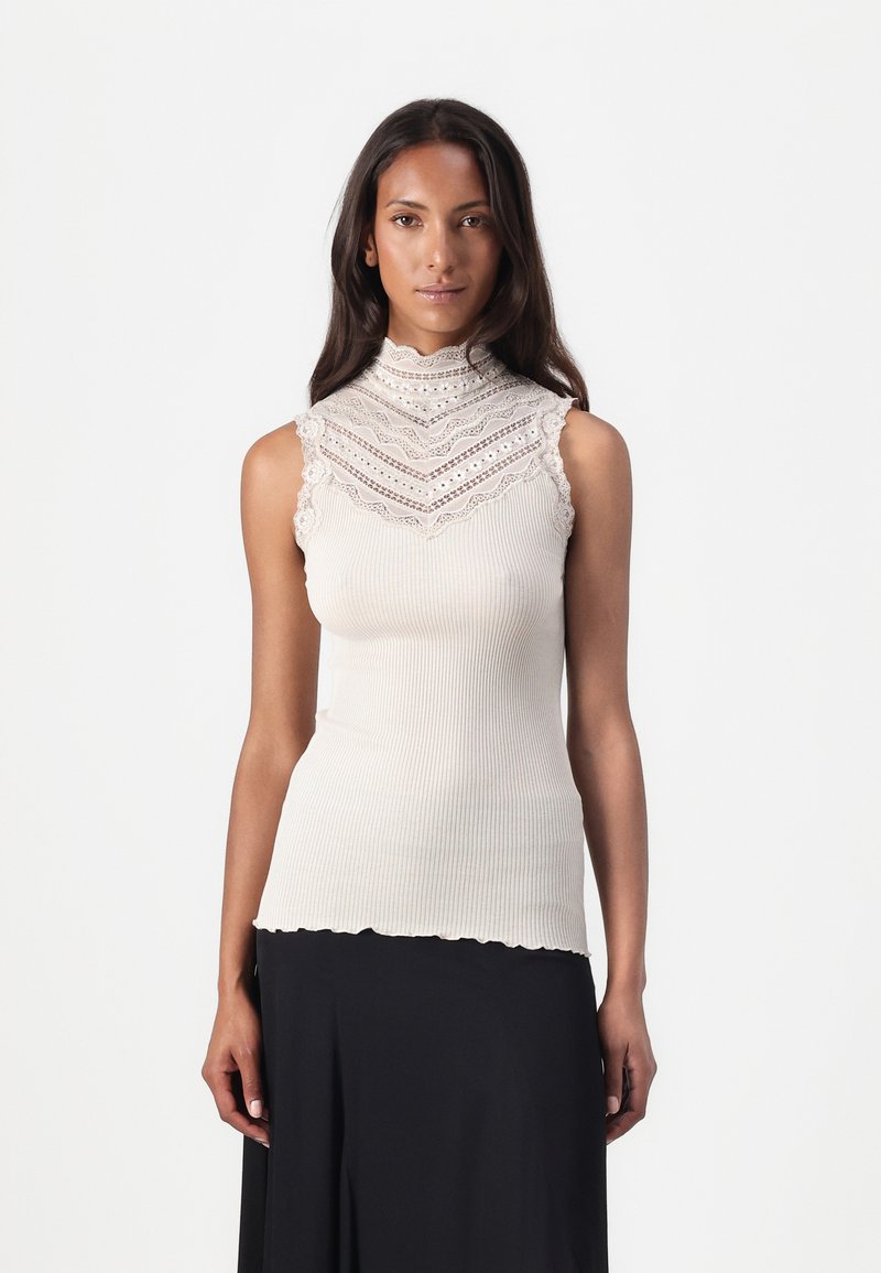 Ribbad ärmlös topp i off-white med hög krage som har intrikata spetsdetaljer och en scalloped nederkant. Slät textur, figurnära design.
