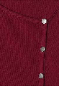 Burgundsko rebrasto blago z okroglim izrezom in petimi srebrnimi kovinskimi pritisnimi gumbi ob levi robu. Mehak material in prilegajoča se zasnova.