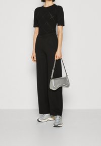 Kvinna i svart kortärmad topp och vida byxor, håller en silvrig handväska, med silvriga och vita träningsskor.