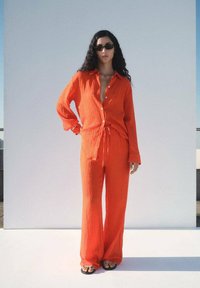 Žiarivý oranžový dvojdielny outfit s košeľou na gombíky a voľnými nohavicami. Tkanina má textúrovaný povrch. Čierne sandále dopĺňajú vzhľad.