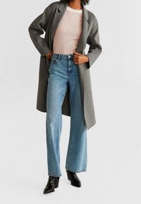 Osoba w szarym długim płaszczu, jasnoróżowym swetrze z dzianiny, wysokich niebieskich szerokich spodniach i czarnych botkach na obcasie.