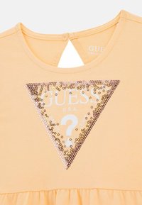 Hellorange T-Shirt mit einem dreieckigen Pailletten-Patch und weißem Text, der "GUESS U.S.A." sowie ein '?' Symbol zeigt. Weicher Stoff mit einem rüschierten Saum.