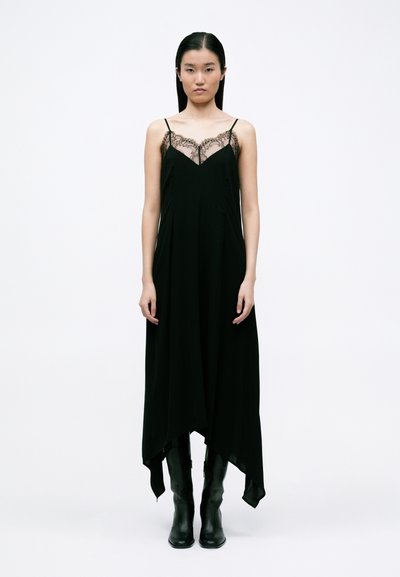 Zwarte maxi-jurk met kantdetails bij de halslijn, verstelbare dunne schouderbandjes en een asymmetrisch zoomontwerp. Model draagt zwarte kniehoge laarzen.