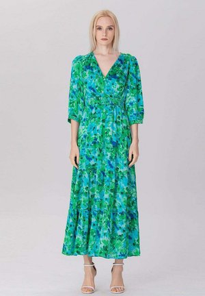 Femme debout face à l'avant, portant une longue robe fleurie verte et bleue avec des manches 3/4 et des talons blancs ouverts au bout, sur un fond uni.