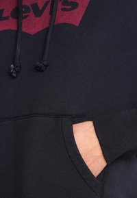 Ruka částečně v přední kapse černé mikiny Levi's s červeným logem a černými šňůrkami.