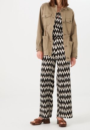 Vrouw draagt een beige jack met knopen over een zwart-wit zigzagpatroon wijd uitlopende broek en bruine sandalen met hak.