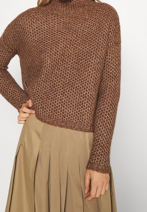 Brun tekstureret strikket sweater båret med høj talje, plisseret beige nederdel, hånd hvilende på hoften, mod en ensfarvet lys baggrund.