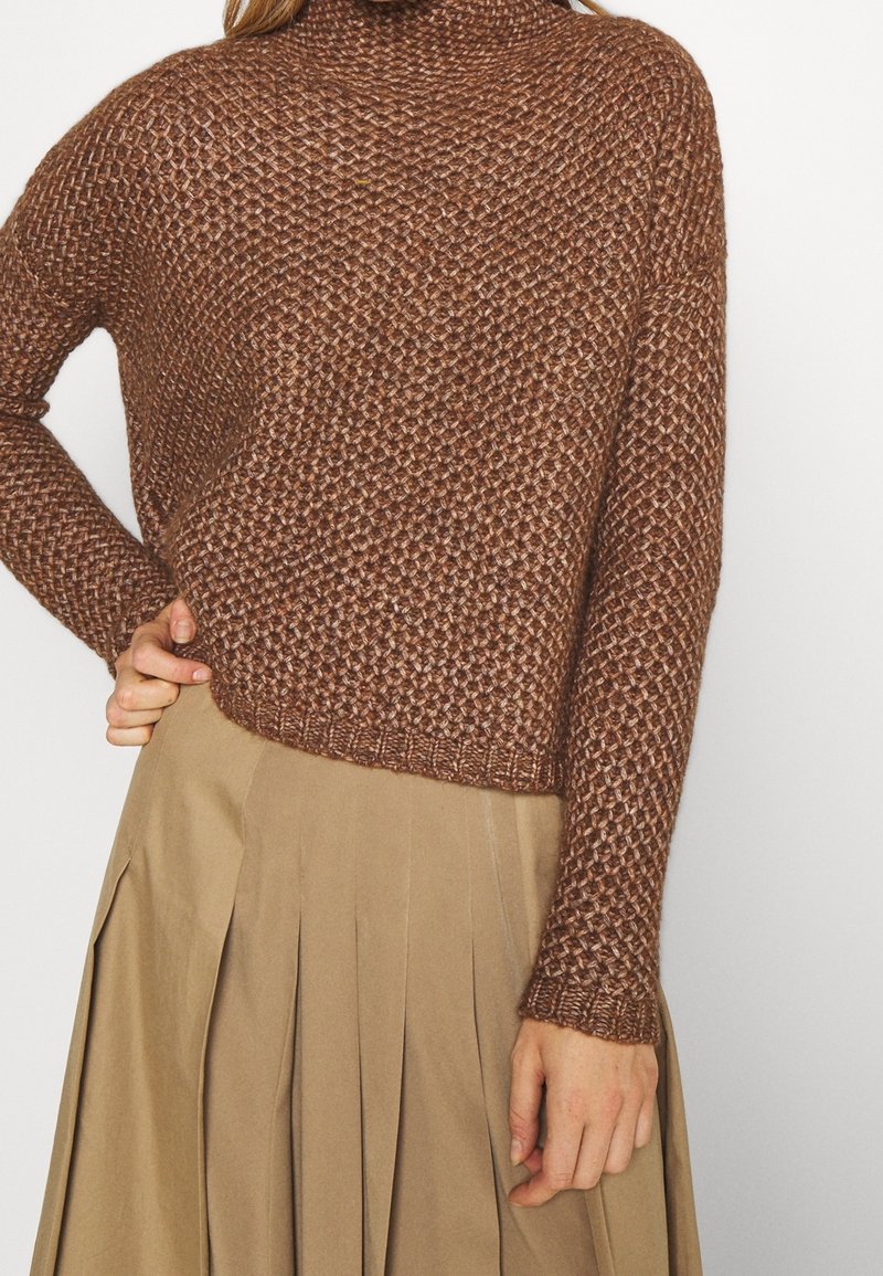 Pull tricoté marron texturé porté avec une jupe beige plissée taille haute, main reposant sur la hanche, sur un fond clair uni.