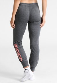 Grijze joggingbroek met een taps toelopende pasvorm, voorzien van een zwart en roze "adidas" logo langs het linkerbeen. Gemaakt van een zachte, gestructureerde stof.