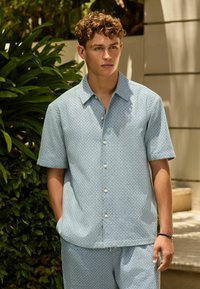 Joven con cabello rizado, vestido con una camisa de manga corta azul claro abotonada y pantalones cortos a juego, de pie al aire libre junto a plantas verdes.