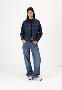 Veste en denim bleu foncé avec des coutures claires, dotée d'un design court, de poches avant et d'une fermeture à boutons. Associée à un jean bleu ample.
