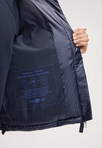 Interno della giacca navy realizzato in tessuto morbido, con etichetta blu che dettaglia il riempimento in poliestere riciclato al 100% e le istruzioni per la cura. Zip nella parte inferiore.