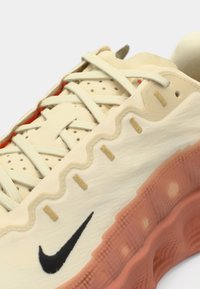Beige athletic shoe with textured upper, contrasting coral sole, and black Nike logo. Features layered design with gold accents and perforations.