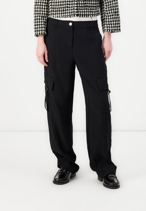 Henkilö, jolla on päällä mustat cargo-housut, joissa on läppätaskut, sekä mustat loafersit, yhdistetty mustavalkoiseen houndstooth-kuvioituun lyhyt takkiin.