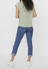 Ljusgrön kortärmad topp med lös passform, kombinerad med blå jeans med upprullade ärmar. Modellen bär vita sneakers och visar en avslappnad outfit från baksidan.