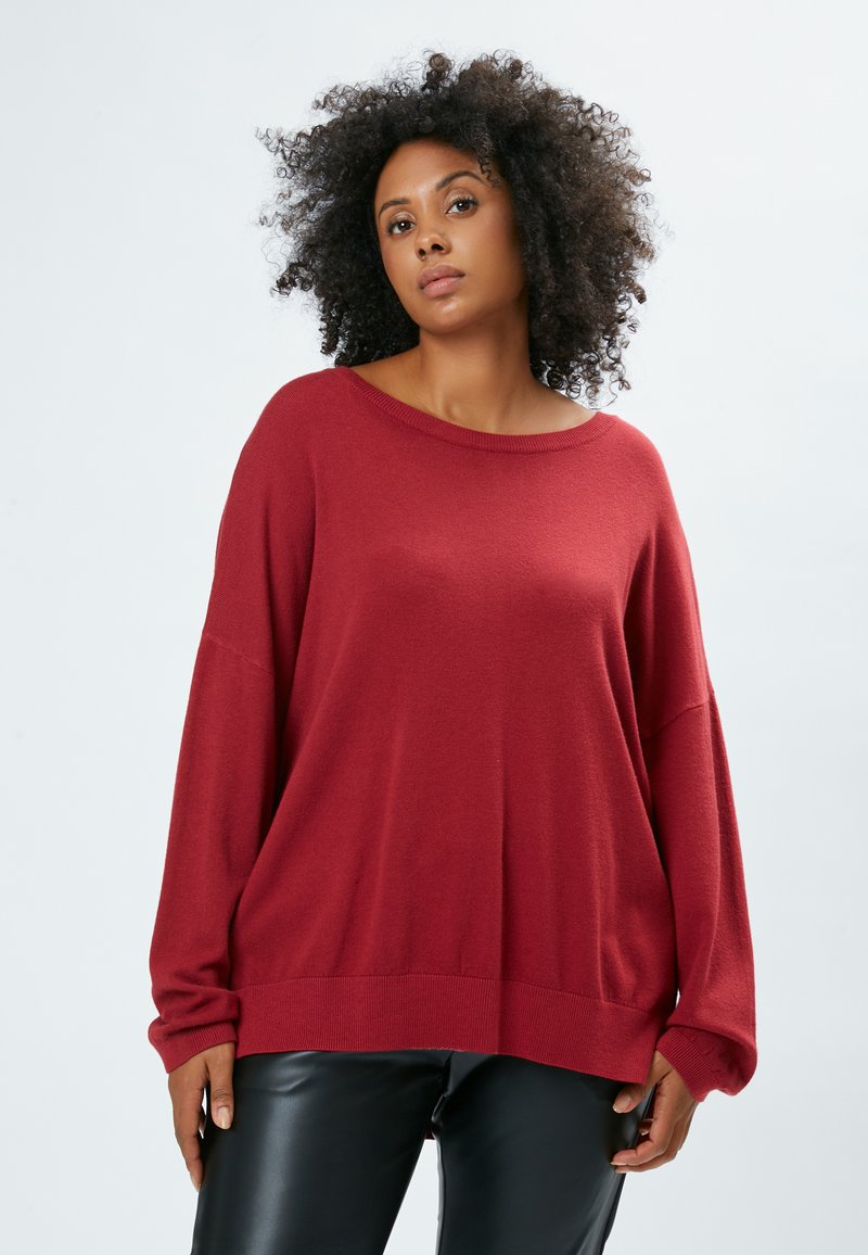 Roter übergroßer Pullover mit rundem Ausschnitt und fallenden Schultern. Weicher Strickstoff und gerippter Saum. Kombiniert mit schwarzen Lederhosen.