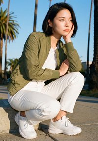 Fiatal nő zöld kabátban és fehér nadrágban guggol a járdán, a fejét a kezére támasztva, mögötte pálmafák.