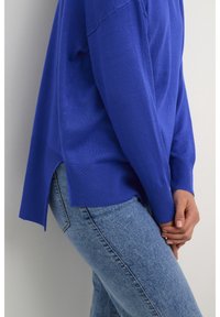Blauer oversized Strickpullover mit gerippten Bündchen, seitlichen Schlitzen und einer weichen Textur, kombiniert mit hellblauen Jeans.