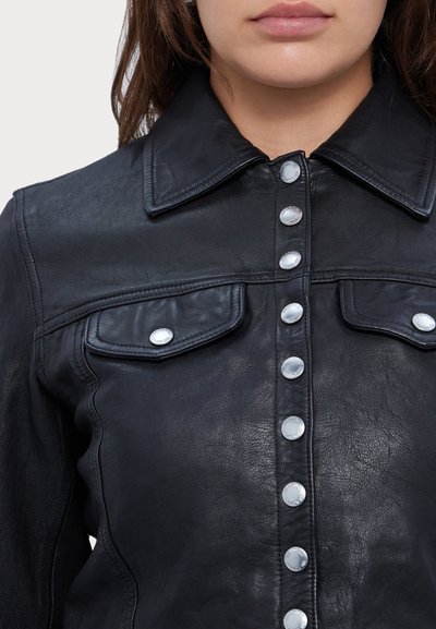 Woman wearing a black leather jacket with silver snap buttons and flap chest pockets, shown from shoulders to mouth.