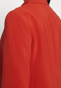 Czerwona marynarka o gładkiej fakturze, z wyraźnie zarysowanymi ramionami i pionowymi szwami, prezentująca dopasowany, nowoczesny krój.