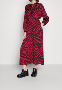 Robe à rayures zébrées rouges et noires, manches longues, taille froncée et encolure en trou de serrure. Portée avec des chaussures à talons compensés noires.