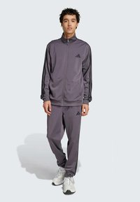 Γκρι αθλητική φόρμα από λεία ύφανση; ζακέτα με φερμουάρ, μαύρες ρίγες και λογότυπο, συνδυασμένη με αντίστοιχες παντελόνες. Άνετη εφαρμογή, χαλαρός σχεδιασμός.