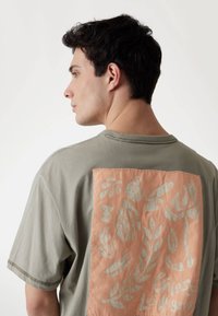 Olivengrøn t-shirt med en koralfarvet applikation på ryggen og lysegrønne botaniske print. Kortærmet og afslappet pasform med synlige sømme.