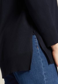 Maglione blu navy a maglia con polsini a coste e spacchi laterali, indossato su jeans in denim azzurro chiaro. Texture liscia, vestibilità casual con spessore medio.