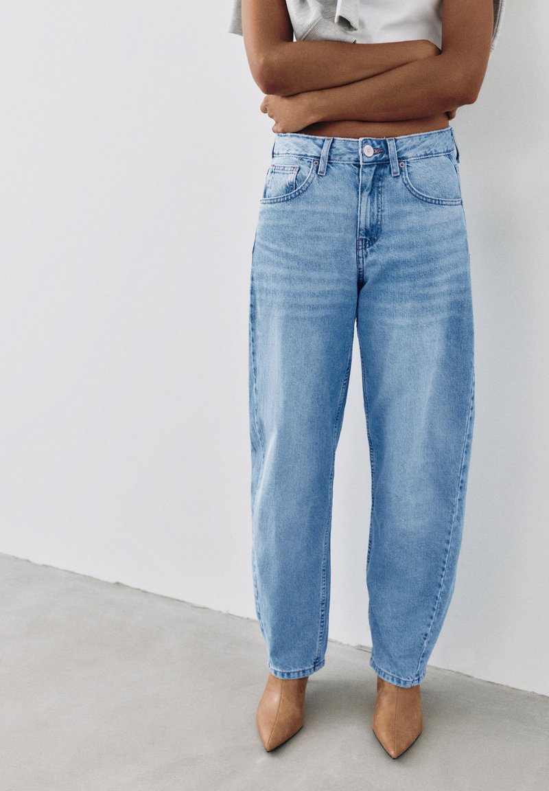 Personne portant un jean taille haute bleu clair et des talons pointus couleur beige, debout, les bras croisés contre un mur blanc.