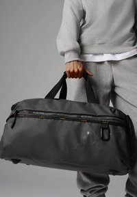 Borsa da viaggio nera a sacca con manici doppi, materiale texturizzato, scomparti con zip e dettaglio del logo. Design compatto e funzionale per i viaggi.