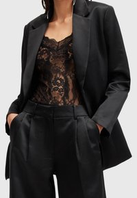 Černý saténový blazer přes průhledný krajkový body. Oblečení obsahuje plisované kalhoty s vysokým pasem, vše v černé barvě, s propracovaným střihem.