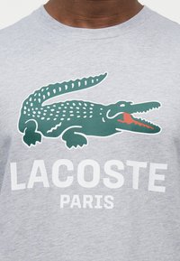 Γκρι πουκάμισο με πράσινο λογότυπο κροκόδειλου πάνω από το έντονο λευκό κείμενο "LACOSTE" και το μικρότερο "PARIS", φορεμένο από ένα άτομο με σκούρο δέρμα.