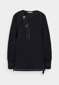 Blouse noire à manches longues avec surpiqûres blanches, une bretelle d'encolure asymétrique et deux boutons décoratifs dorés à l'avant.