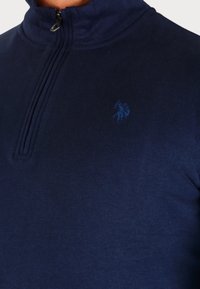 Marineblauwe pullover met een hoge kraag, voorzien van een half-rits sluiting en een subtiel tonale logo op de borst, gemaakt van zachte stof.