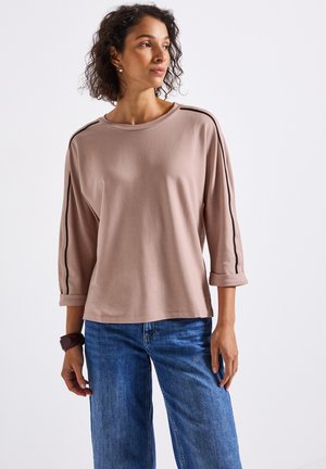 Femme avec des cheveux bouclés portant un tee-shirt mauve à manches longues avec des rayures noires sur les côtés et un jean bleu, se tenant devant un fond blanc uni.