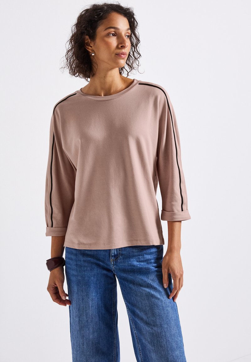 Frau mit lockigem Haar, die ein mauvefarbenes Langarmshirt mit schwarzen Seitenstreifen und blaue Jeans trägt, vor einem schlichten weißen Hintergrund stehend.