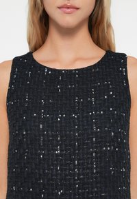 Gros plan sur une femme portant un haut noir sans manches en tissu texturé avec de petites paillettes réfléchissantes dispersées sur le tissu.