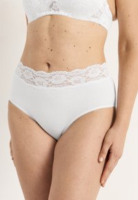 Nainen pukeutuneena valkoisiin puuvillaisiin alusvaatteisiin, joissa on pitsinen vyötärönauha, esittää keskivartalon ja käsi lepää reisellä.