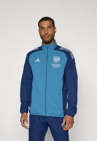 Blau und navy Zip-Jacke mit Adidas-Streifen, Arsenal-Logo und dem Text "Emirates Fly Better". Hergestellt aus leichtem, glattem Stoff.