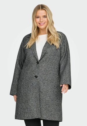 Mujer sonriente con cabello rubio largo que lleva un abrigo de lana gris de gran tamaño sobre una camiseta blanca, de pie frente a un fondo claro y liso.