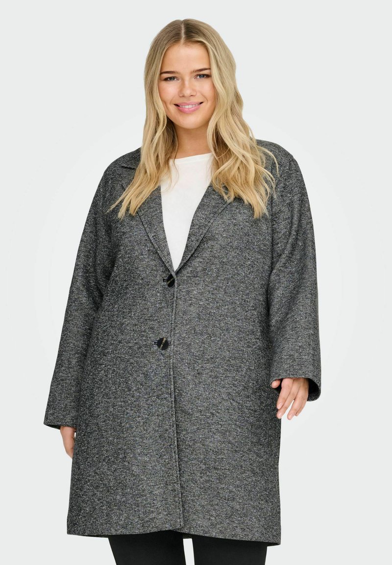 Femme souriante aux longs cheveux blonds portant un manteau en laine gris oversize par-dessus un haut blanc, debout devant un fond clair uni.