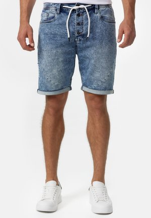 Mand iført oppbrettede blå denimshorts med sorte knapper og hvid snor, parret med hvide sneakers på en hvid baggrund.