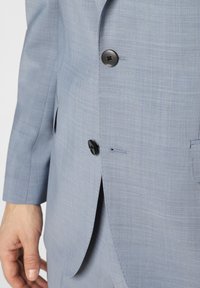 Jasnoniebieska marynarka garniturowa o gładkiej fakturze, z pojedynczym czarnym guzikiem do zapięcia i widocznym przeszyciem na krawędzi. Posiada boczne rozcięcie.