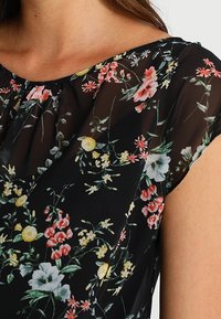 Kwiatowa bluzka wykonana z czarnego, przezroczystego materiału, z żywymi, wielokolorowymi wzorami kwiatowymi. Posiada zaokrąglony dekolt i krótkie rękawy.