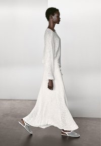 Biały dziergany top z długim rękawem i szerokimi rękawami, zestawiony z zwiewną białą maxi spódnicą oraz szarymi butami wsuwanymi. Gładka tekstura na całej powierzchni.