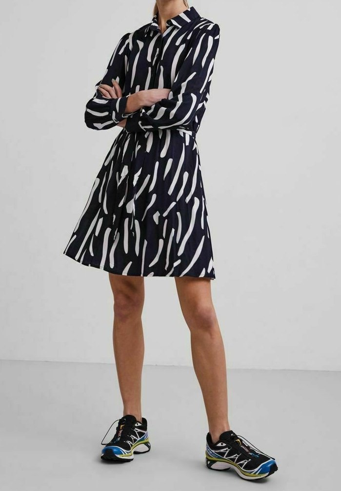 Donna in piedi con le braccia incrociate, indossa un vestito a maniche lunghe con motivo bianco e nero e scarpe sportive nere, blu e gialle, contro uno sfondo semplice.