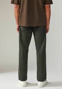 Pantalons vert olive à coupe droite, avec deux poches arrière à rabats inclinés, associés à un t-shirt marron et des baskets blanches.
